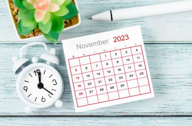 November 2023 Monthly calendar year and alarm clock with pen on blue wooden background.