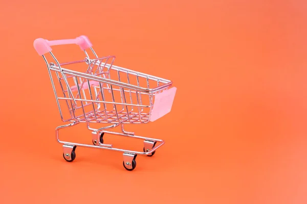 Small supermarket grocery push cart for shopping toy with wheels isolated on orange pastel colorful paper trendy background.