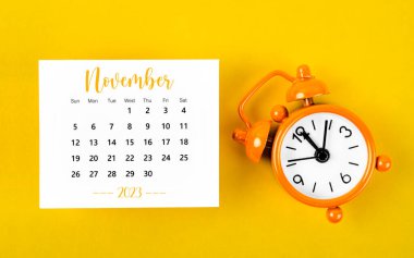 2023 Kasım ayının takvimi. Sarı arka planda siyah alarm saati var..