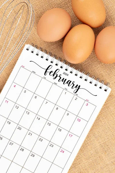 stock image February 2024 Desk calendar and fresh eggs with egg whisk on burlap background.