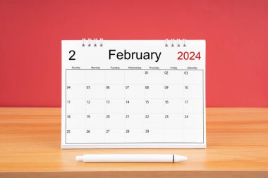 2024 yılı için Şubat ayının masa takvimi ve kırmızı arkaplan ile ahşap masa üzerinde kalem.
