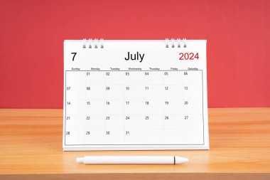 2024 yılı için Temmuz aylık masa takvimi ve kırmızı arkaplan ile ahşap masa üzerinde kalem.