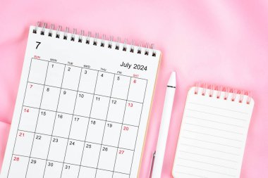 Temmuz 2024 takvim ve pembe kumaş arka planında açık günlüğü olan kalem. Aylık takvim kavramı.