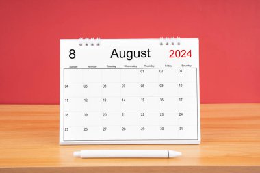 2024 yılı için Ağustos ayının masa takvimi ve kırmızı arkaplan ile ahşap masa üzerinde kalem.