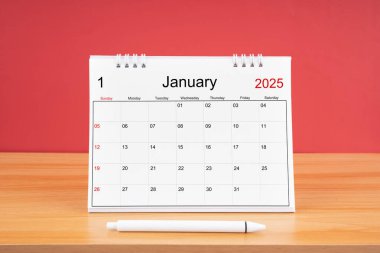 2025 yılı için Ocak ayının masa takvimi ve kırmızı arkaplan ile ahşap masa üzerinde kalem.