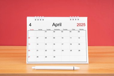 2025 yılı için Nisan aylık masa takvimi ve kırmızı arkaplan ile ahşap masa üzerinde kalem.