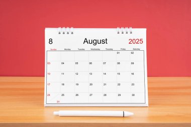 2025 yılı için Ağustos ayının masa takvimi ve kırmızı arkaplan ile ahşap masa üzerinde kalem.