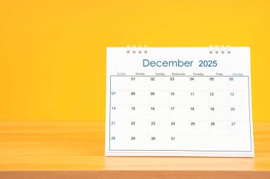 Aralık 2025, 2025 yılı için aylık masa takvimi sarı arka planlı ahşap masa üzerinde.