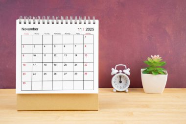 2025 yılı için Kasım Aylık masa takvimi ve kırmızı arkaplan ile ahşap masa üzerindeki alarm saati.