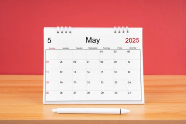 2025 yılı için aylık masa takvimi ve kırmızı arkaplan ile ahşap masa üzerinde kalem.