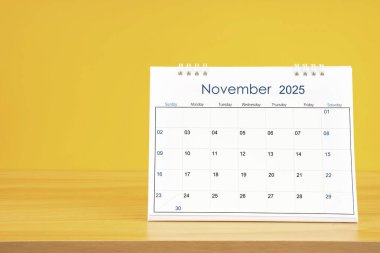 Kasım 2025, 2025 yılı için aylık masa takvimi sarı arka planlı ahşap masa üzerinde.
