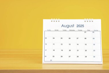Ağustos 2025, 2025 tarihli aylık masa takvimi sarı arka planlı ahşap masa üzerinde..