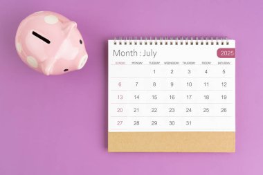 Temmuz 2025. Masa takvimi ve mor arka planda pembe kumbara..