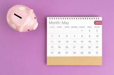 Mayıs 2025 Masa Takvimi ve pembe kumbara mor arka planda.