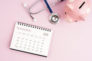 Kasım 2025. Masa takvimi ve domuz kumbarasıyla steteskop, sağlık konseptleri için tasarruf.