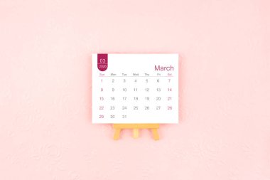 Pembe arka planda ahşap sehpası olan Mart 2026 takvim sayfası.