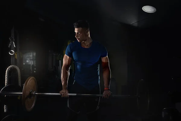一个男人在体育馆里举重 穿着紧身T恤衫 带着创造性的照片朝旁边看去 一些强壮的运动健将锻炼身体的概念背景 肌肉健美的健美男子赤身裸体地在体育馆里锻炼 — 图库照片