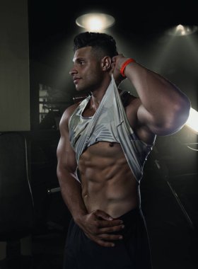 Vücut geliştirici spor salonunda karın kaslarını gösteriyor. Tişörtünü kaldırıyor, yaratıcı bir fotoğraf çekiyor. Dar elbiseli seksi bir adam.