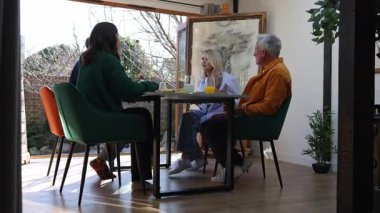 Aile üyeleri bir masada oturuyor, kahvaltıdan zevk alıyor ve sohbet ediyorlar.