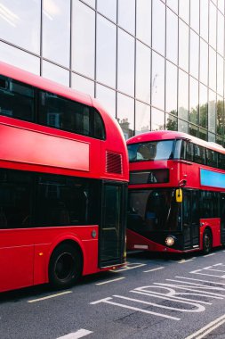 İki kırmızı çift katlı otobüs Londra caddesinde park halinde, modern bir ofis binasının cam cephesine yansıyor.