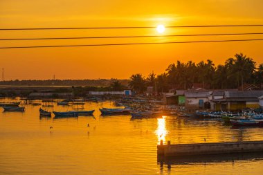 Ilık ışıkta parlayan turuncu bir gün batımı ve sakin suların altın gökyüzünü yansıttığı sakin bir kayıkhane. Balıkçı tekneleri kıyıya yakın sularda huzur içinde dinleniyor.