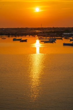 Ilık ışıkta parlayan turuncu bir gün batımı ve sakin suların altın gökyüzünü yansıttığı sakin bir kayıkhane. Balıkçı tekneleri kıyıya yakın sularda huzur içinde dinleniyor.