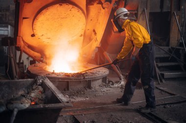 Bir metal işçisi endüstriyel üretim sırasında fırında erimiş metal karıştırıyor.