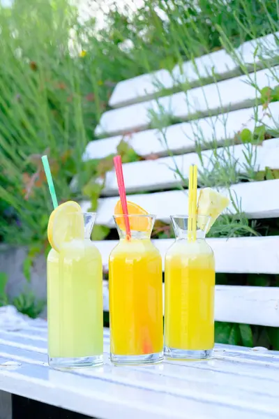 Açık havada narenciye dilimleriyle süslenmiş cam şişelerdeki üç taze meyve suyu.