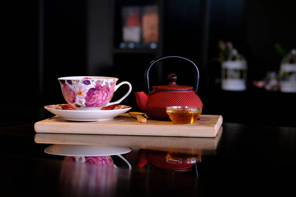 Floral teacup and red teapot set on a wooden board reflecting on a dark surface