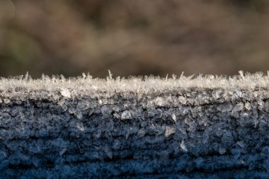 Buz kristalleri nazikçe aşınmış ahşap bir yüzeye tutunarak yaklaşırken buz oluşumlarının karmaşık ayrıntıları bulanık arka plana vurgulanır.