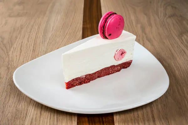 Beyaz kremalı bir dilim kırmızı kadife pasta ve ahşap bir masada beyaz bir tabakta pembe bir makaron.