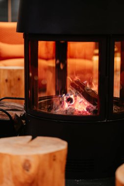 A close-up of a modern fireplace with glowing embers and a partially burned log. Wooden log tables and a textured orange couch are visible in the background.
