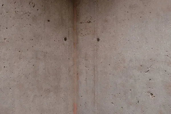 A close-up of a concrete wall shows a raw, unadorned surface marked by subtle variations in color, scattered small holes, and a corner construction detail.
