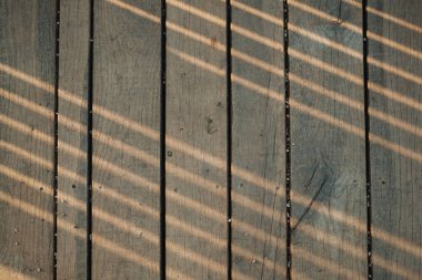 Güneş ışığı, yıpranmış ahşap bir güvertede doğrusal gölgeler oluşturur. Bu model dinamik dokular ve soyut estetik cazibe yaratır..