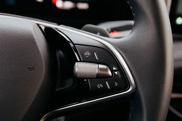 Bir arabanın direksiyon simidinin odaklanmış görüntüsü, çeşitli kontrol düğmeleri ve dokulu bir tutuş. Teknolojiyi ve rahatlığı vurgulamak. Karanlık iç dekorasyon.