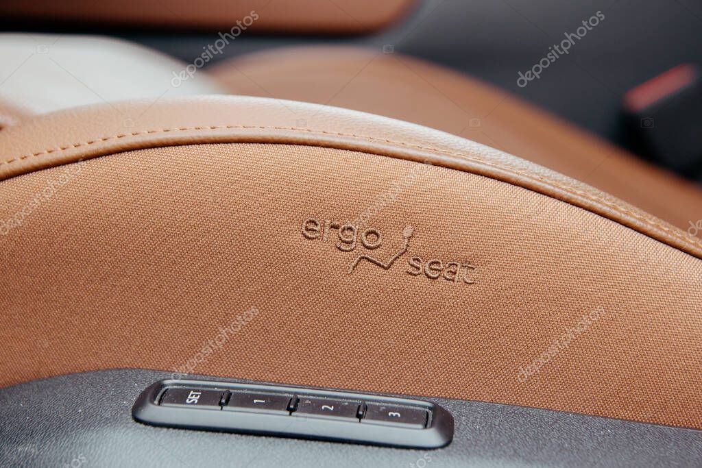 Close-up of a car seat featuring lumbar support and adjustment settings. The seat is upholstered in light brown fabric with an embroidered logo. Black trim.