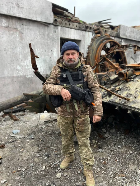 An elderly Ukrainian soldier stands against background of a broken rusty tank with torn off turret