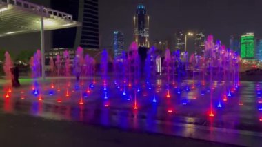 Aydınlatmalı açık havuz. Farklı renklerde su, değişen yoğunluk ve akış yönü. Arka planda bir grup insan gece su gösterisini izliyor..