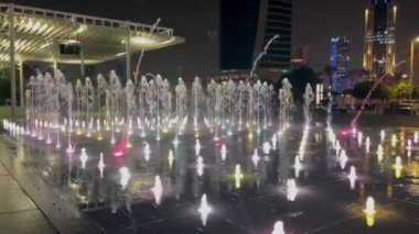 Aydınlatmalı açık havuz. Farklı renklerde su, değişen yoğunluk ve akış yönü. Arka planda bir grup insan gece su gösterisini izliyor..