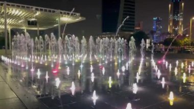 Aydınlatmalı açık havuz. Farklı renklerde su, değişen yoğunluk ve akış yönü. Arka planda bir grup insan gece su gösterisini izliyor..