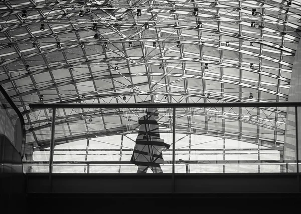 A man is walking by a big glass and steel ceiling. His shadow is reflected to the glass railing.