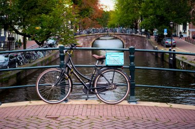 Tek bir bisiklet, Amsterdam ikonik kanalı ve kemerli köprüler tarafından mükemmel çerçevelenmiş korkuluklara dayanır. Bu sessiz kompozisyon şehir ritminin özünü yakalar: sakin, güzel ve her zaman bisikletle hareket eden.