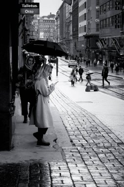 Genç bir kadın binanın duvarında yağmurun durmasını bekliyor. Diğer insanlar şemsiyelerle onun yanından geçiyorlar..
