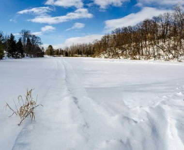 Donmuş kar birikintisi mavi bulutlu gökyüzünün altında kar motosikleti izleri ile kaplı.