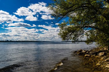 St. Lawrence Nehri, dalgalı kıyı şeridi, mavi gökyüzü, güneşli bulutlar ve ön planda kayalık kıyı şeridi