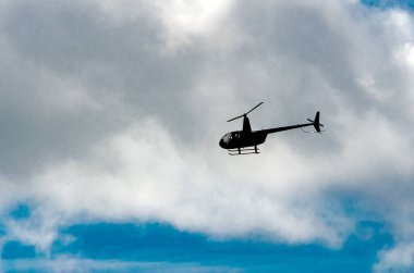 Mavi gökyüzünün üstündeki bulutların üzerinde sola uçan bir helikopterin siluetli yan görüntüsü.