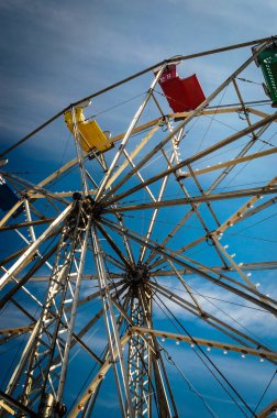 Antika bir dönme dolabın altındaki mavi gökyüzüne karşı birincil renkli koltuklar ve metal yapı..