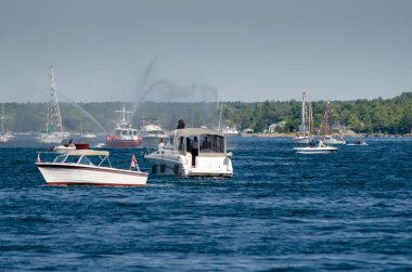 İtfaiye teknesi St. Lawrence Nehri 'ndeki bir yelkenliye su püskürtüyor. Arka planda New York eyalet kıyı şeridi var..
