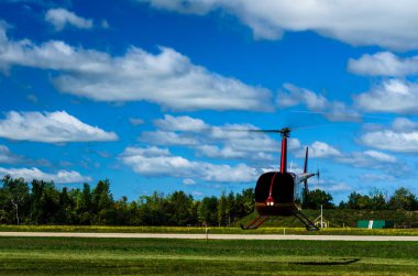 Arka planda bulutlu mavi bir gökyüzü ve bir ağaç çizgisi ile pistte süzülen helikopter..