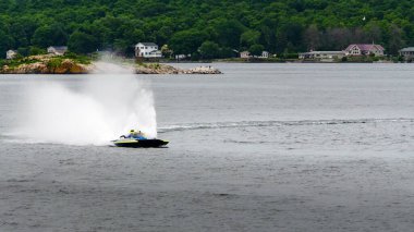 Arka planda bir ada ve sahil kulübeleriyle birlikte sağa dönen mavi ve sarı yarış teknesi sprey püskürtüyor.